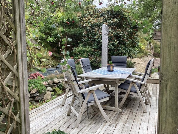 A wooden deck with a table and chairs, surrounded by greenery and a small stream. - 4 Star Holiday Home in Munkedal (Munkedal)