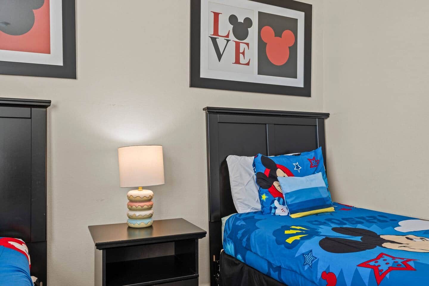 A hotel room with a bed featuring Mickey Mouse-themed bedding, a nightstand with a lamp, and two framed pictures on the wall.