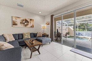 A modern living room with a sectional sofa, a coffee table, and a glass door leading to an outdoor area with a pool. - Serenity Resort | Private Pool Townhome | 3Bd3bath (Four Corners)