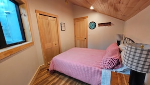 Ein Schlafzimmer mit Holzboden, einem Bett mit karierter Bettwäsche, einem Fenster und einer Lampe auf einem Nachttisch.