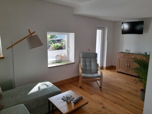 Una sala de estar con un sofá, una silla, una mesa de centro con un control remoto, una ventana con vistas y un televisor en la pared.