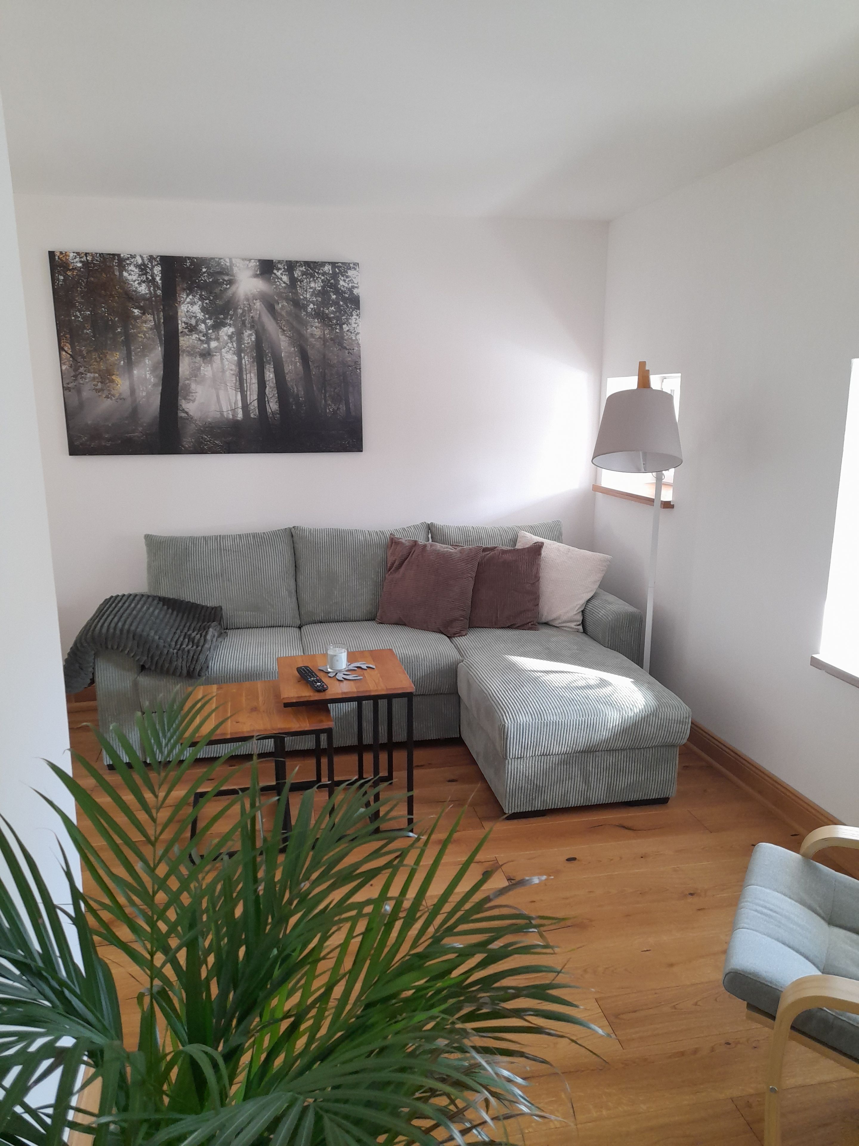 Ein Wohnzimmer mit einem Ecksofa, einem Couchtisch, einer Stehlampe und einem großen Wandbild mit Waldmotiv.
