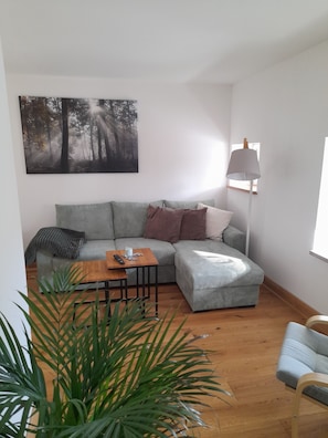 Un salon comprenant un canapé d’angle, une table basse, un lampadaire et un grand tableau représentant une forêt, accroché au mur.