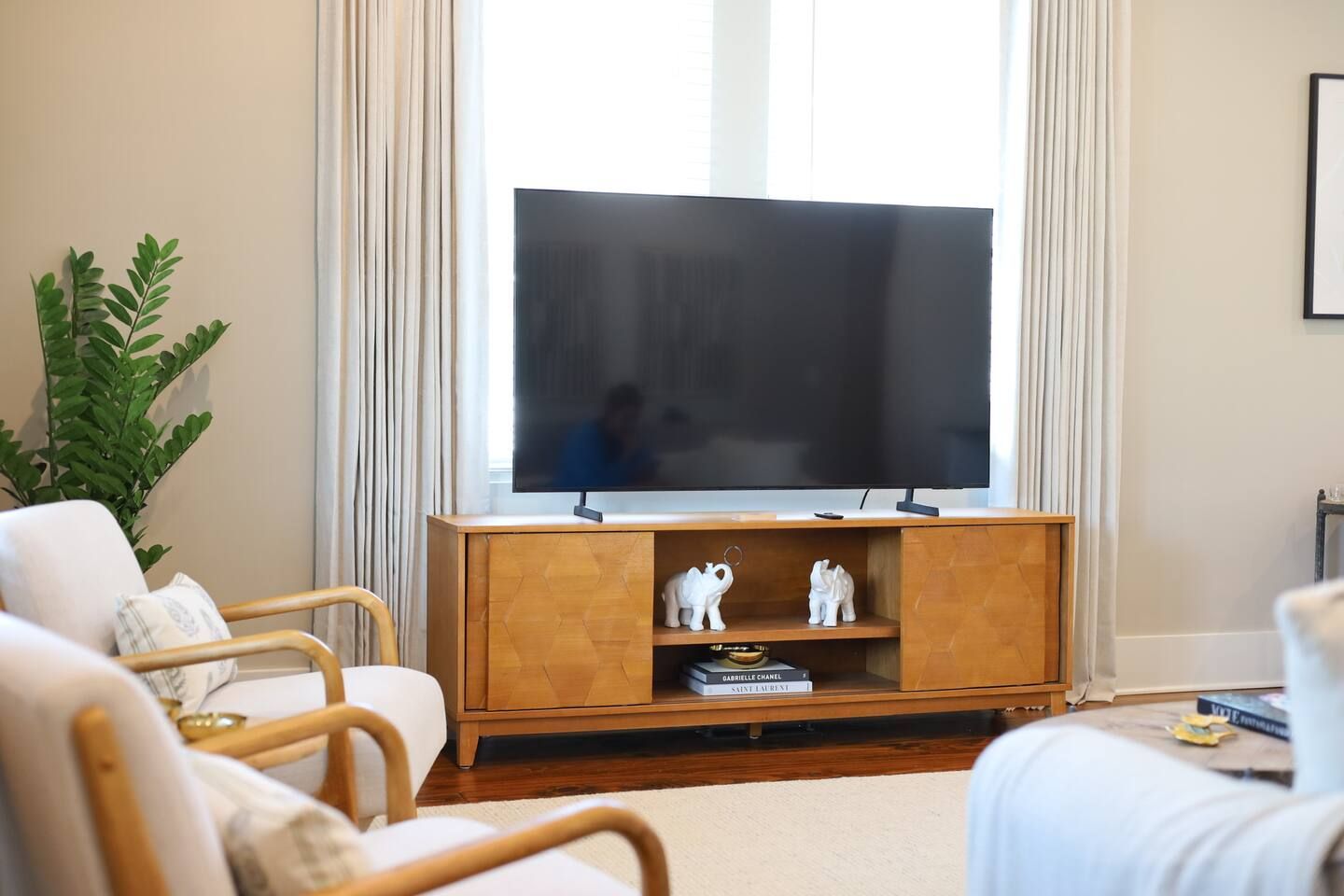 Ein Wohnzimmer mit einem Flachbildfernseher auf einem Holzständer, zwei weißen Elefantfiguren, einer Topfpflanze und einem Sofa mit Kissen.