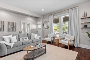 A modern living room with a grey sofa, two armchairs, a coffee table, and a large window with curtains.