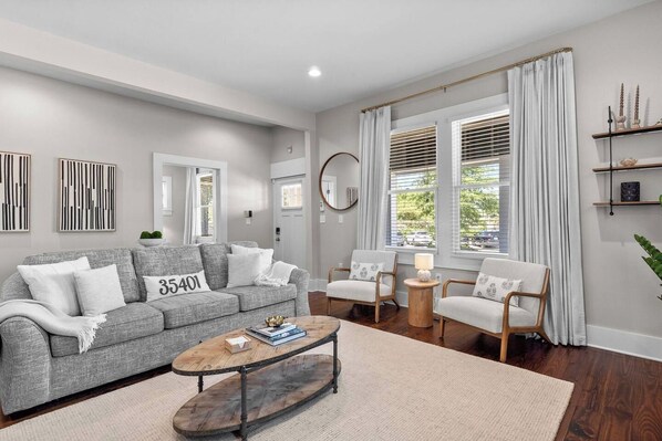 Una sala de estar moderna con un sofá gris, dos sillones, una mesa de centro y un ventanal con cortinas.