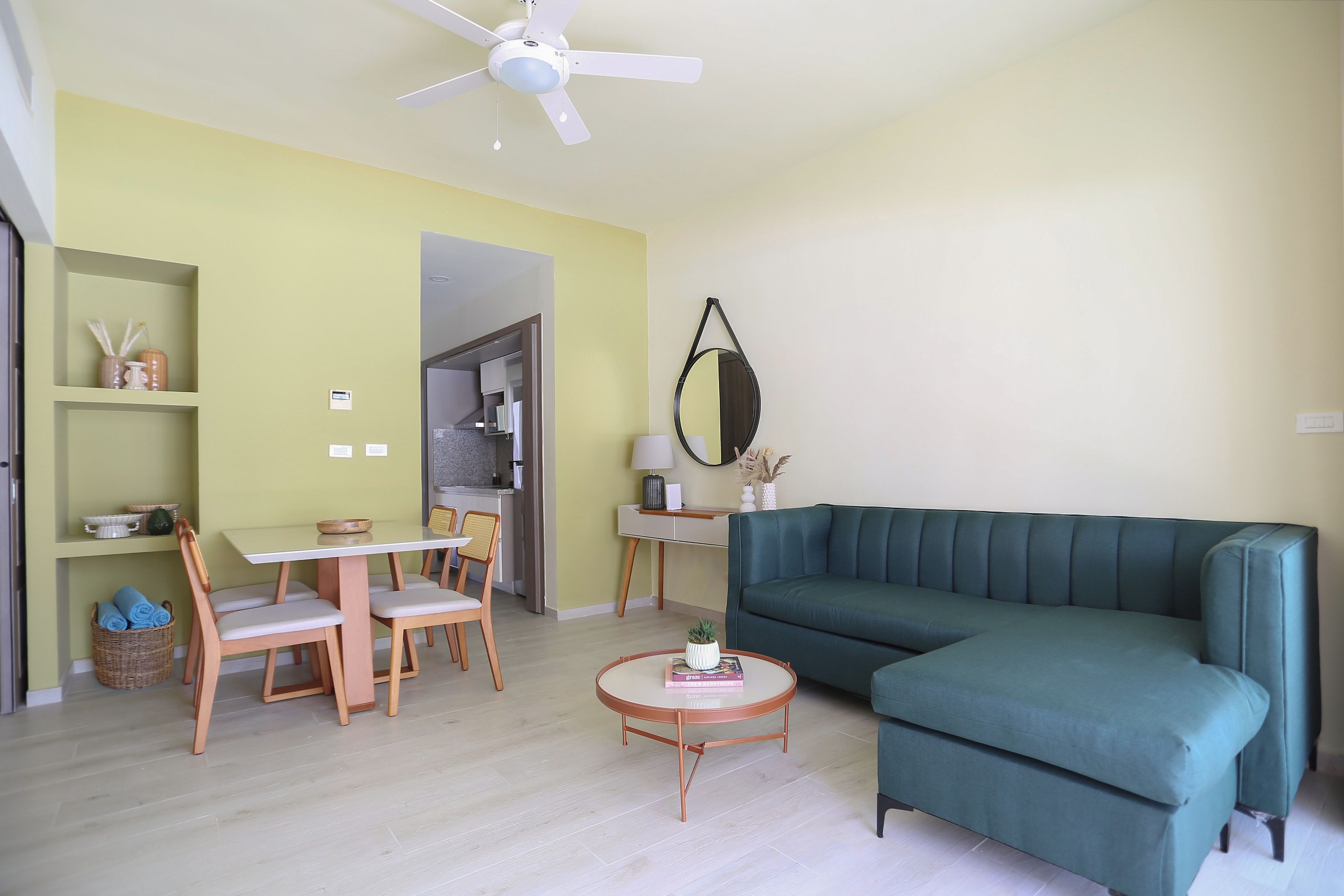 Una sala de estar con un sofá verde, una mesa de comedor con sillas, un ventilador de techo y una cocina al fondo.