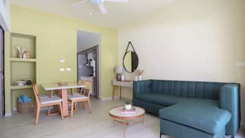 Un salon avec un canapé vert, une table à manger avec des chaises, un ventilateur de plafond et, au fond, un coin cuisine.