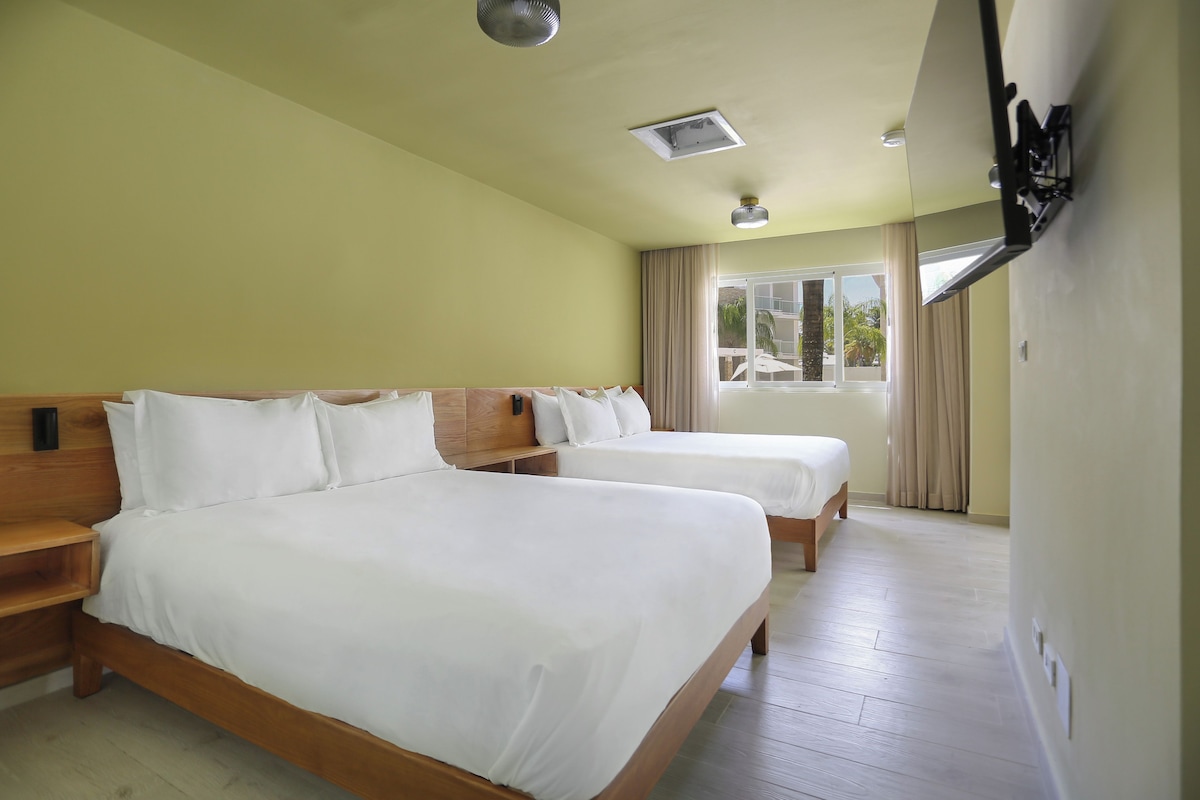 Habitación de hotel con dos camas, cabeceros de madera, una ventana con cortinas y un ventilador de techo.