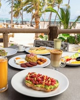 A table set with breakfast items including eggs, toast, and a beverage, with a view of palm trees and the sea in the background.
