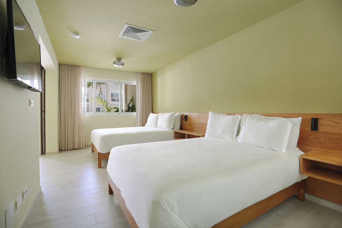 Habitación de hotel con dos camas, una ventana con cortinas, un respiradero en el techo y un cabecero de madera.