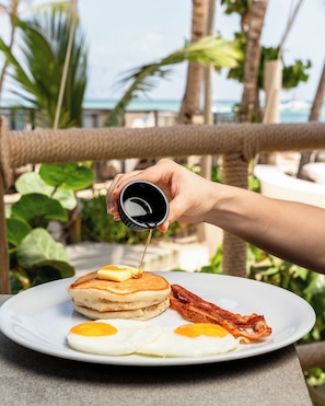 A breakfast plate with pancakes, eggs, and bacon, with syrup being poured on top.