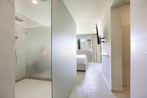 A modern bathroom with a glass shower enclosure, a visible toilet, and a bed in the adjacent room.
