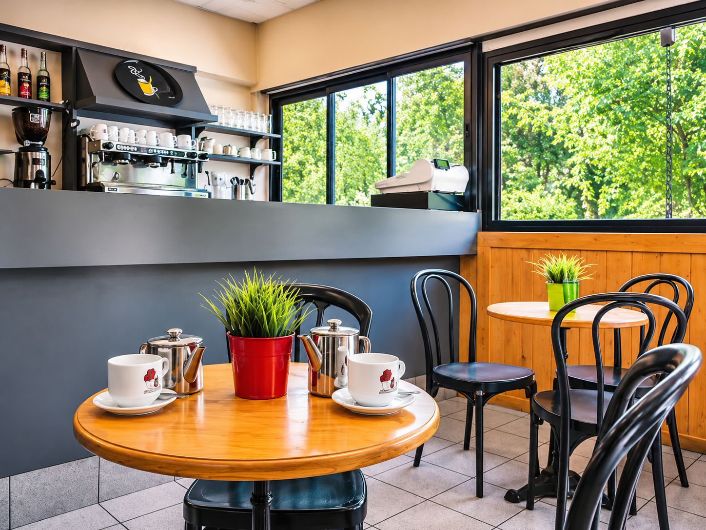 Een café met een koffiemachine, kopjes en uitzicht op groen.