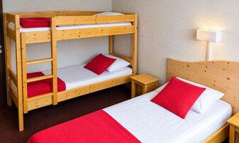 Una cama litera con dos niveles, una cama individual con almohada roja, una mesita de noche de madera y una lámpara fijada a la pared.
