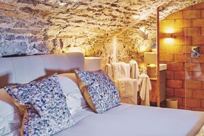 Ein Schlafzimmer mit einer Steinstur, einem Bett mit floralen Kissen, einem Nachttisch mit Lampe und einer Ziegelwand mit Spiegel.