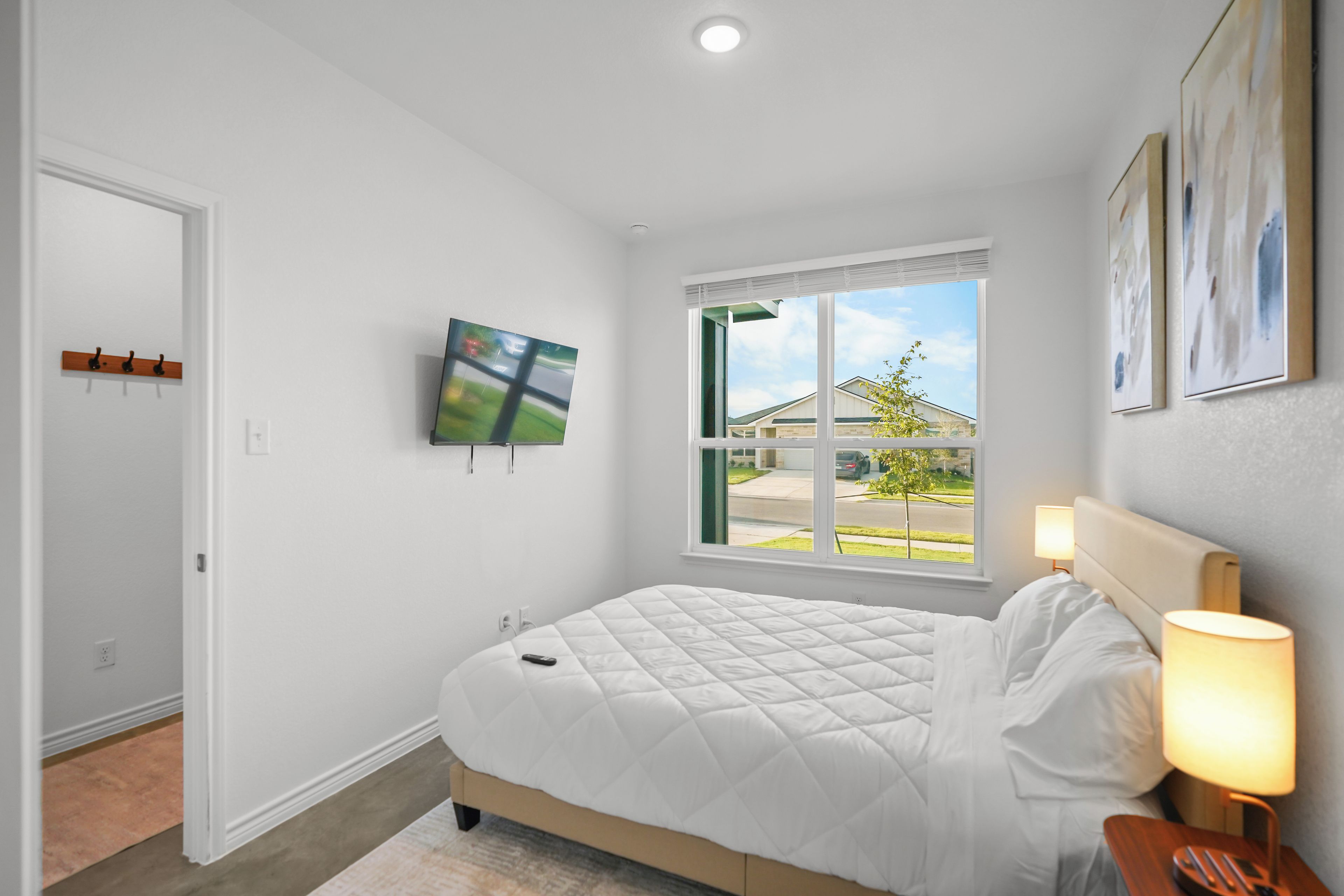 A bedroom with a bed, a TV mounted on the wall, a window with a view of the outside, and a lamp on the bedside table.