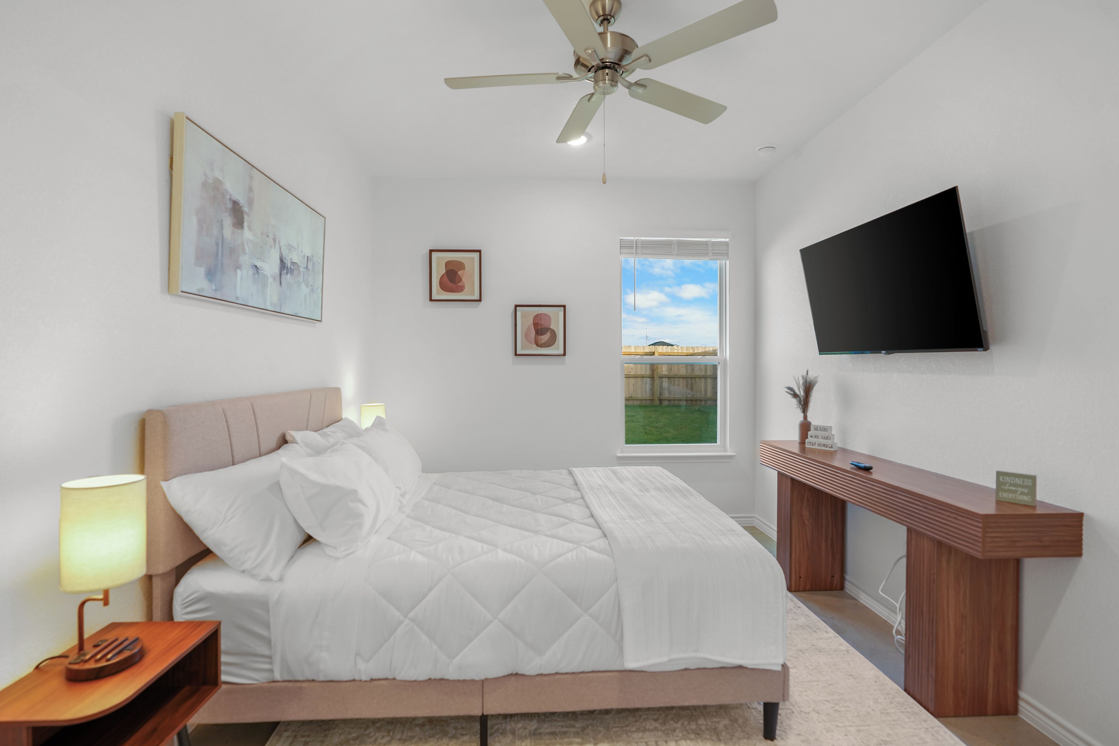 A bedroom with a bed, a bedside table with a lamp, a ceiling fan, a TV mounted on the wall, and artwork on the walls.