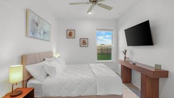A bedroom with a bed, a bedside table with a lamp, a ceiling fan, a TV mounted on the wall, and artwork on the walls.