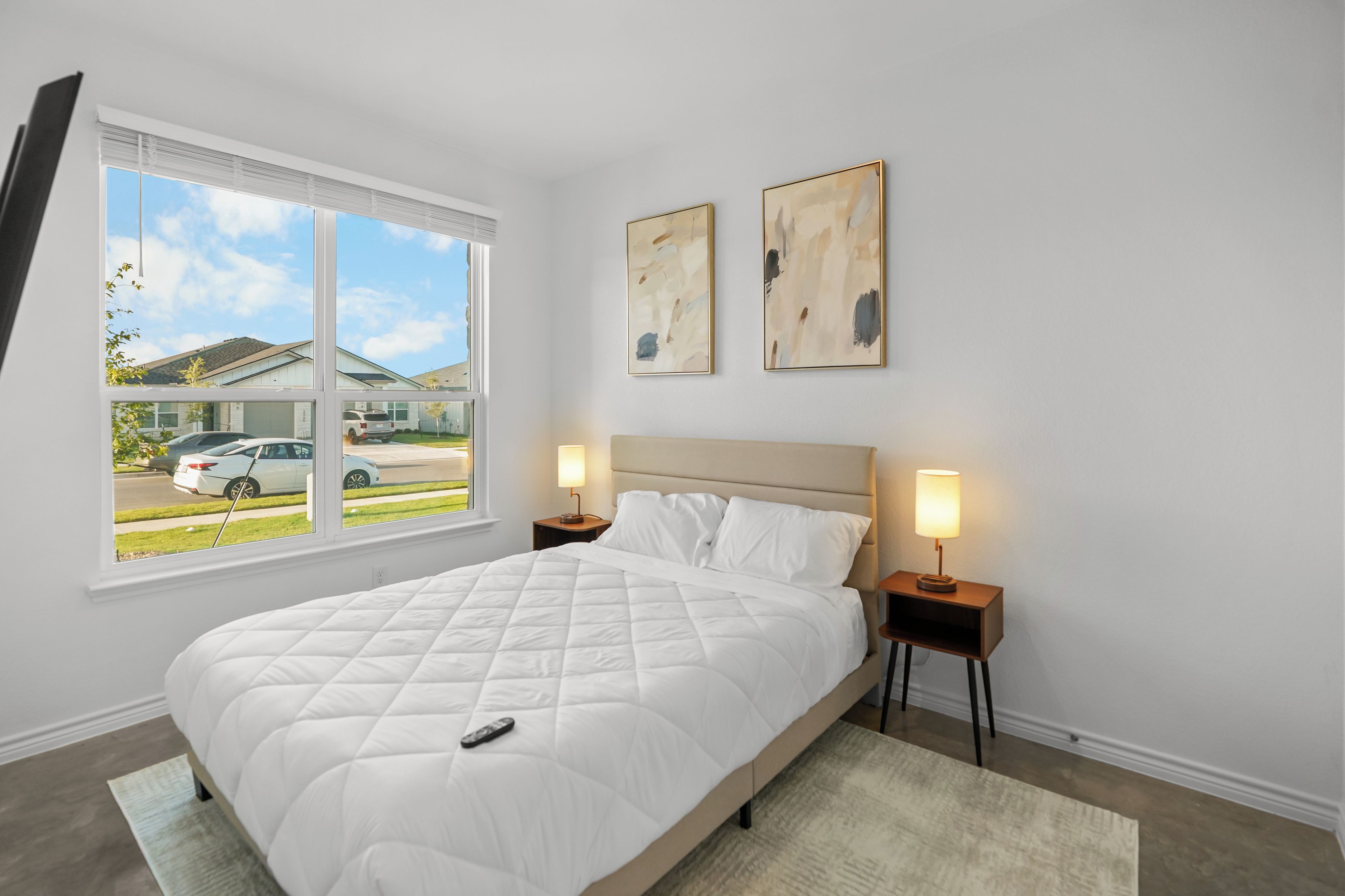 A bedroom with a bed, two bedside lamps, a window with a view of parked cars, and two abstract paintings on the wall.