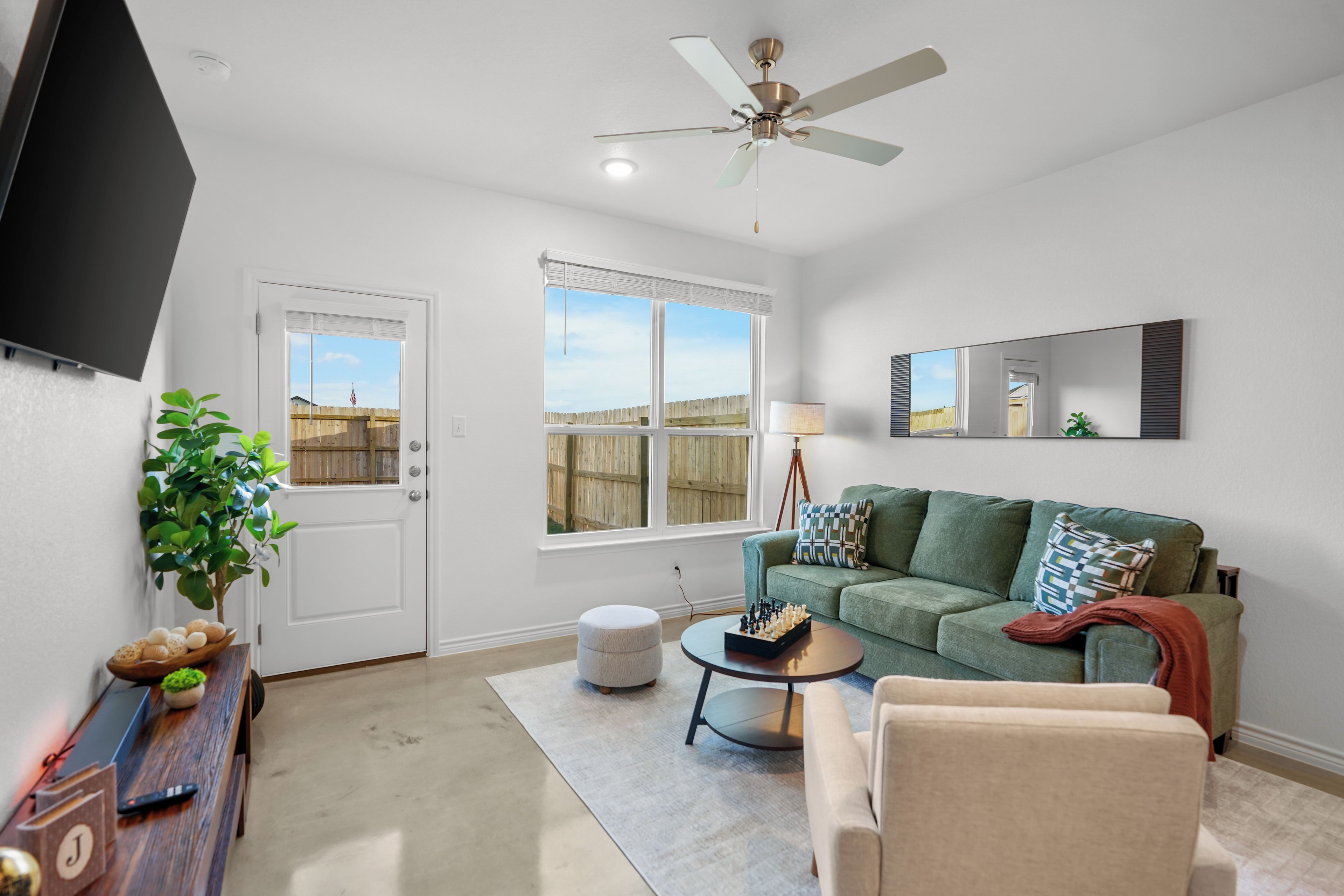 A living room with a green sofa, a coffee table, a TV mounted on the wall, a ceiling fan, and a window with a view of a wooden fence.