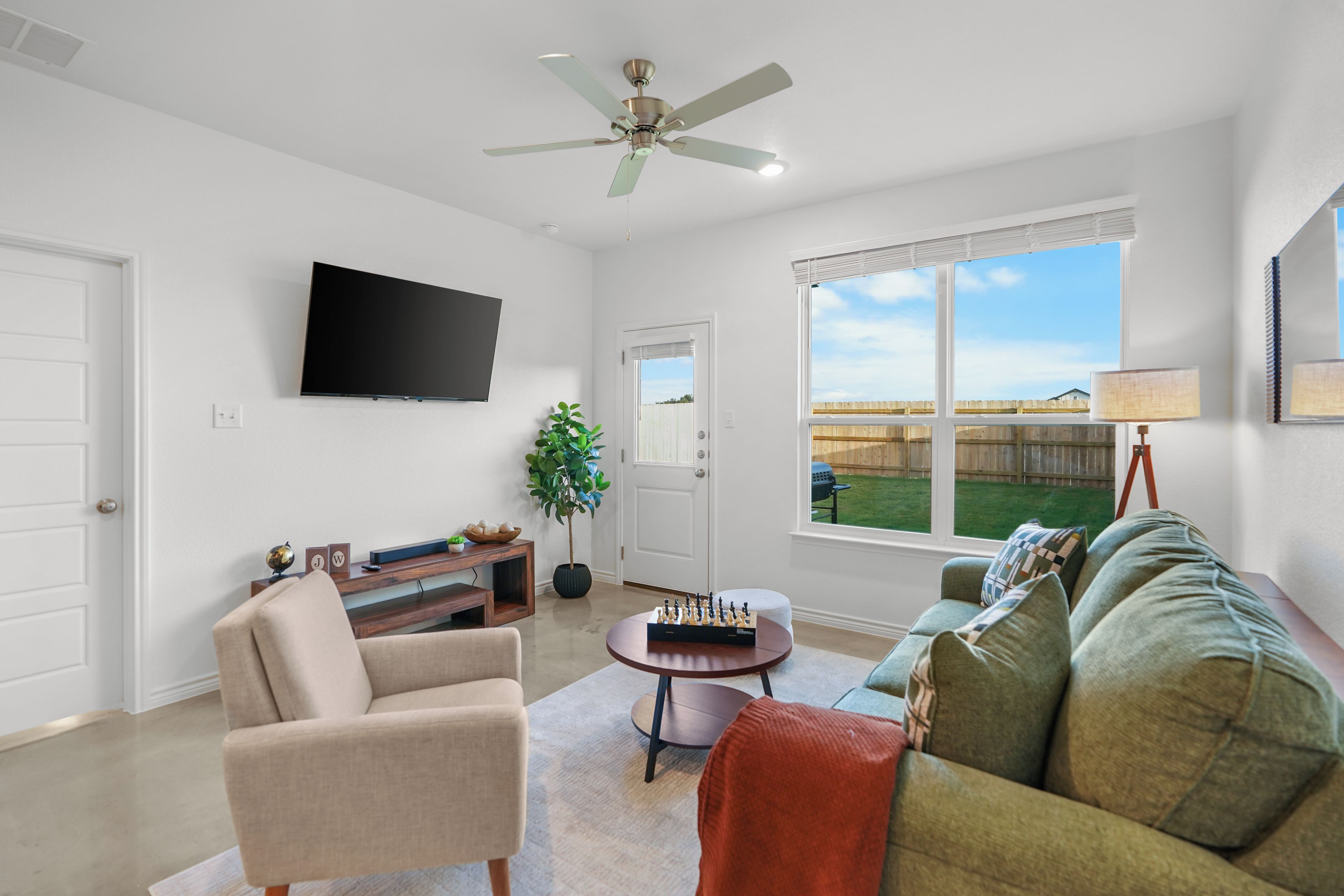 A living room with a flat-screen TV, a ceiling fan, a large window with a view of a backyard, and a green couch.
