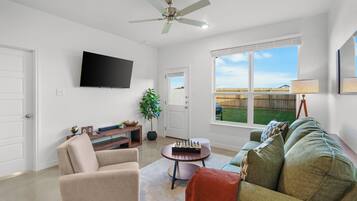A living room with a flat-screen TV, a ceiling fan, a large window with a view of a backyard, and a green couch.