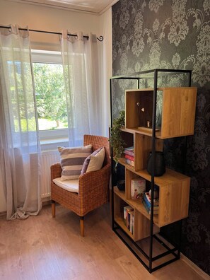 A room with a wicker chair, a bookshelf, and a window with sheer curtains. - Ilztalperle – A Modern Getaway Right on the Ilz River in the Bavarian Forest (Hutthurm)