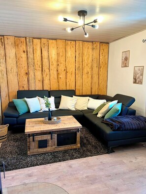 A modern living room with a sectional sofa, a wooden coffee table, and a ceiling light fixture. - Ilztalperle – A Modern Getaway Right on the Ilz River in the Bavarian Forest (Hutthurm)