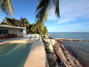 A poolside area with a clear view of the ocean, palm trees, and a building with outdoor furniture. - A genuine Yucatecan Summer House by the Sea (Chicxulub)