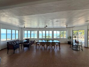 En rummelig stue med et stort spisebord, flere stole og en sofa. Stuen har store vinduer med udsigt til havet.