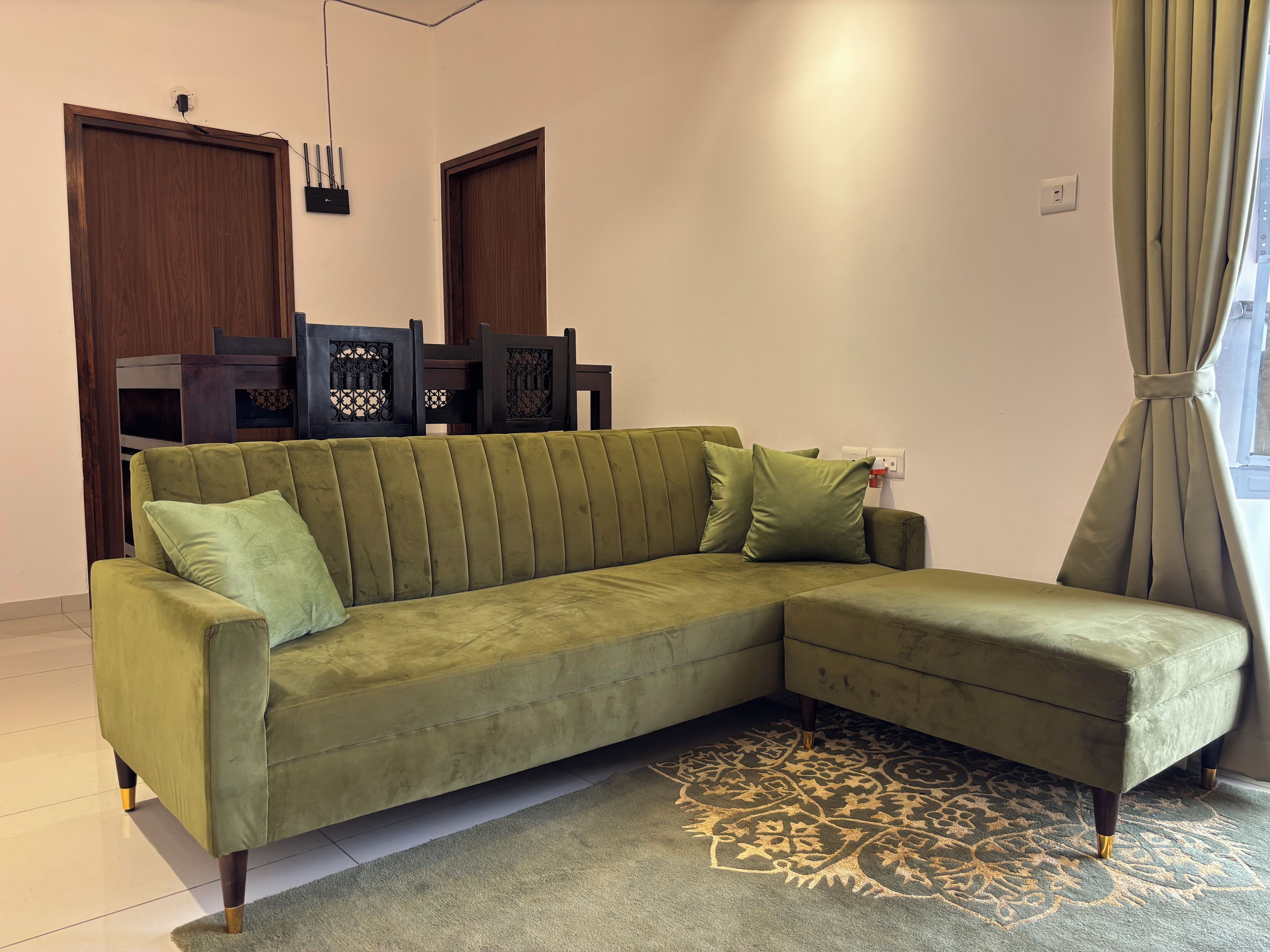 A green sofa with a patterned rug, wooden chairs, and a window with curtains.