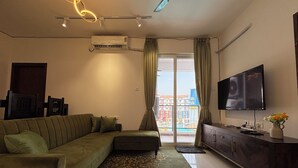 Una sala de estar con un sofá modular, un televisor de pantalla plana, un aire acondicionado moderno y vistas a edificios de colores a través del ventanal.