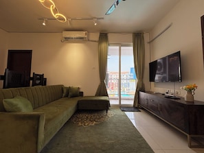 A living room with a sectional sofa, a flat-screen TV, a modern air conditioner, and a view of colorful buildings through the window. - Spacious 3-bedroom apartment with AC in gorgeous Chicalim (Chicalim)