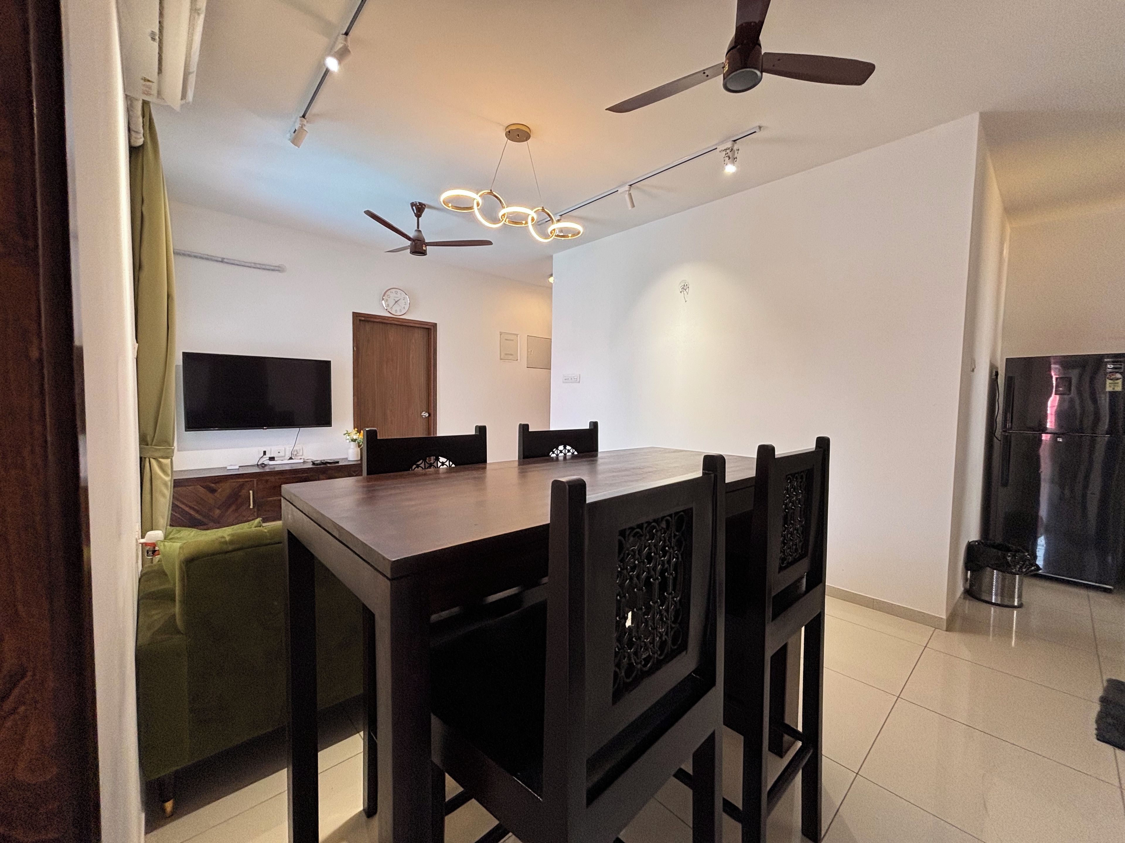 A modern dining area with a rectangular table, six chairs, a ceiling fan, recessed lighting, a flat-screen TV, a wooden cabinet, and a refrigerator.