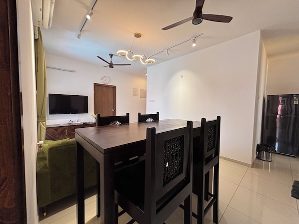A modern dining area with a rectangular table, six chairs, a ceiling fan, recessed lighting, a flat-screen TV, a wooden cabinet, and a refrigerator.