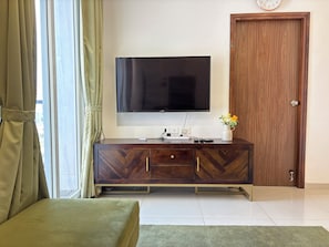 A room with a large flat-screen TV mounted on the wall, a wooden cabinet with drawers, a green upholstered bench, and a wooden door.