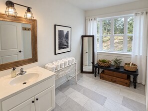 A bathroom with a white sink, a mirror, a framed picture, towels, and a window with a view of trees.