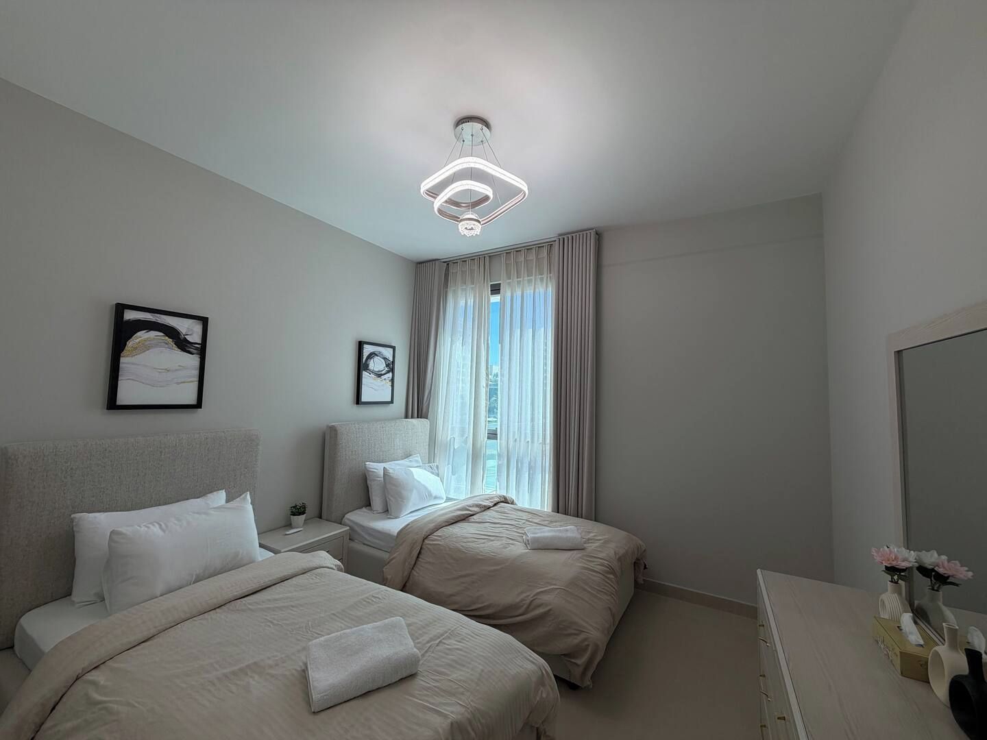 Une chambre d’hôtel avec deux lits, une table de chevet, une lampe de chevet, une grande fenêtre avec des rideaux et un luminaire au plafond.