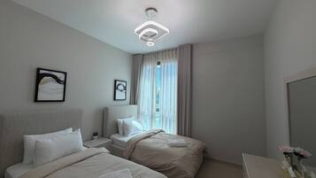 Une chambre d’hôtel avec deux lits, une table de chevet, une lampe de chevet, une grande fenêtre avec des rideaux et un luminaire au plafond.