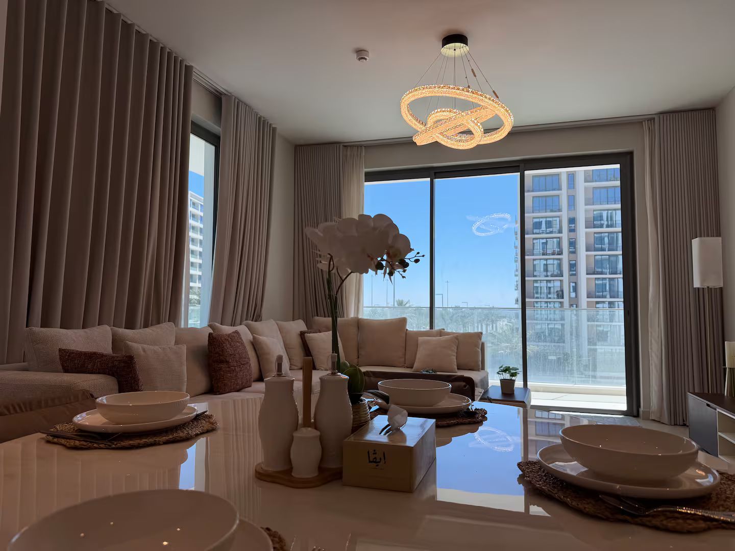 A modern living room with a large window, a sofa set, a coffee table with decorative items, and a unique chandelier.