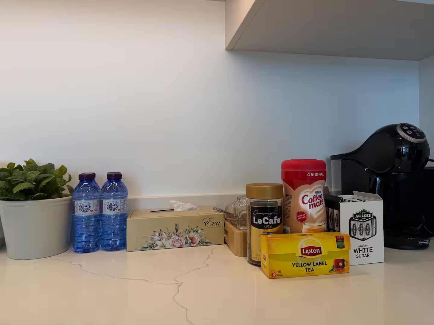 Een keukenblad met diverse items, waaronder flessen water, een doos Lipton thee, een potje LeCafe, een koffiebus en een koffiezetapparaat.