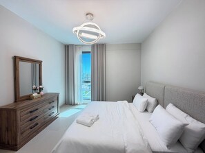 A modern bedroom with a large bed, a wooden dresser, a mirror, and a view of the outdoors. - Executive 2BR Stay in Marassi Terrace FMT202O (محافظة المحرق)