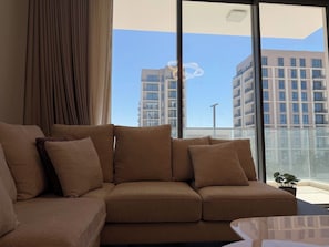 A modern living room with a sofa and a view of high-rise buildings through large windows. - Executive 2BR Stay in Marassi Terrace FMT202O (محافظة المحرق)