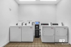 A laundry room with two washing machines, a dryer, and a control panel. - Golden Sunsets - 2 Bed, Pool, Beach, Fun (Gulf Shores)