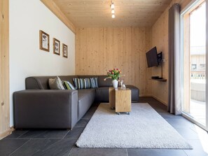 A modern living room with a grey sectional sofa, a small wooden side table with a vase of flowers, and a flat-screen TV mounted on the wall. - One-bedroom Cabin (Sankt Georgen am Kreischberg)