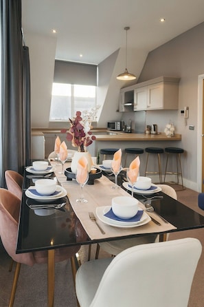 A dining area with a long table set for six, a kitchen in the background, and a modern interior design. - The Stylish Artist Penthouse | Central Harrogate (Harrogate)