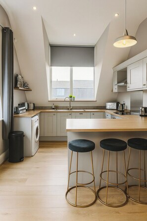 A modern kitchen with a central island, bar stools, a washing machine, and a window with a view. - The Stylish Artist Penthouse | Central Harrogate (Harrogate)