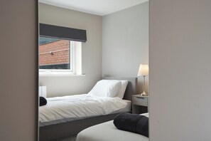 Een kleine kamer met een enkel bed, een raam, een nachtkastje met lamp en uitzicht op een gebouw van baksteen.