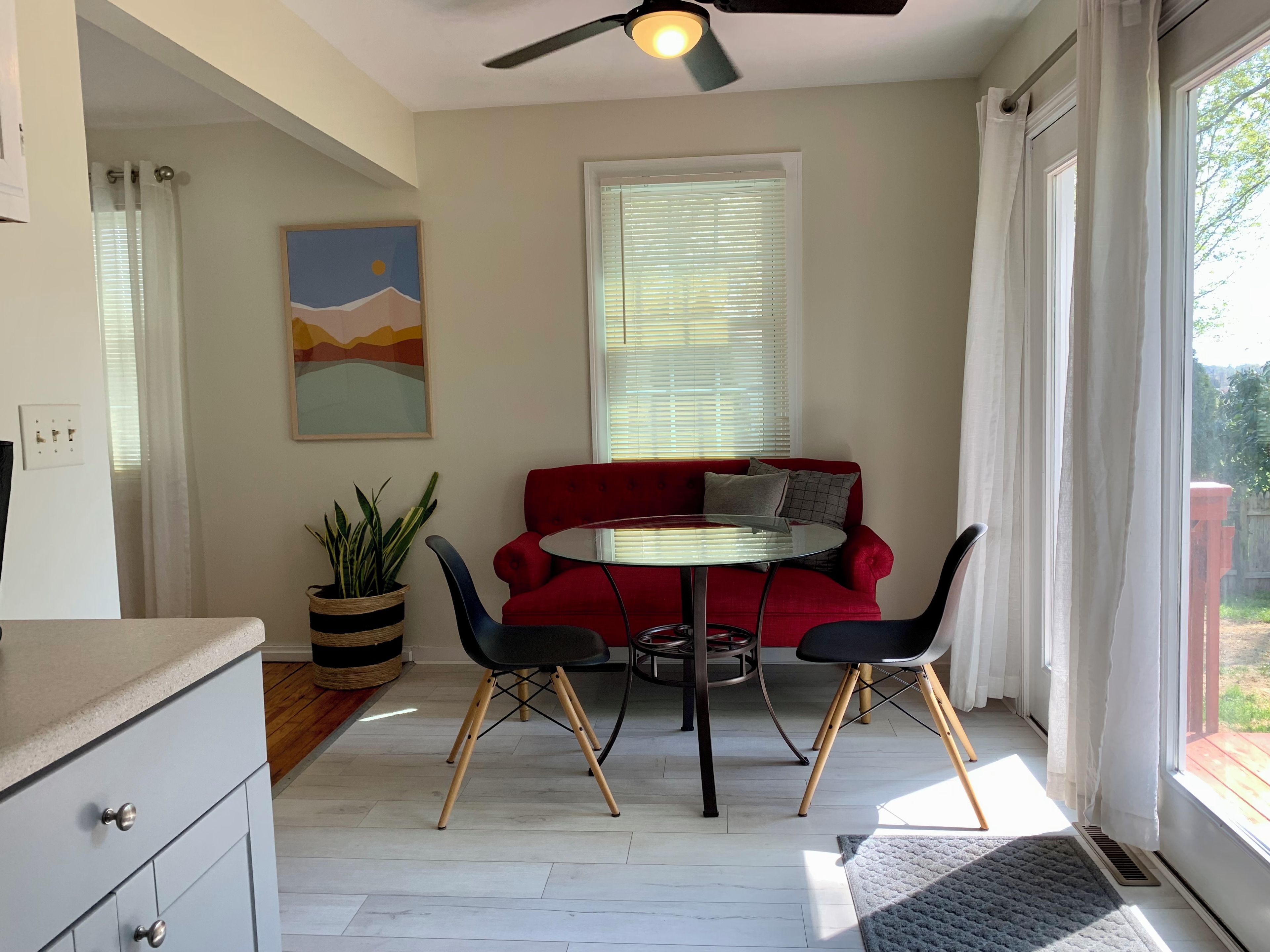 Una habitación con un sofá rojo, una mesa redonda y dos sillas. Hay un ventilador de techo, una ventana con persianas y un cuadro en la pared.